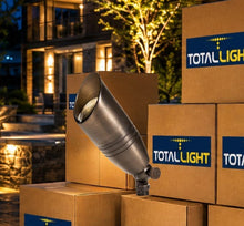Load image into Gallery viewer, A TotalLight outdoor landscape light in a brushed metal finish, shown resting on top of stacked cardboard boxes with the brand logo visible on the boxes. The background is a softly blurred evening outdoor setting with a house and warm string lights.