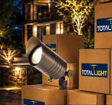 Load image into Gallery viewer, A TotalLight outdoor landscape spotlight in a dark metallic finish, shown illuminated and angled, with several branded cardboard boxes stacked around it in a nighttime garden setting.
