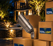 Load image into Gallery viewer, A brushed metal outdoor light fixture mounted on a stack of brown cardboard boxes, with the TOTALLIGHT logo visible on the boxes, set against a softly blurred nighttime residential yard with illuminated trees and a house.
