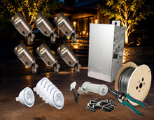 Load image into Gallery viewer, An outdoor landscape lighting kit displayed on a wooden deck at night, featuring six silver spotlight heads, a silver metal control box, a spool of wire, white downlight bulbs, wire connectors, and green-handled crimping tools, with a warm-lit house in the background.

